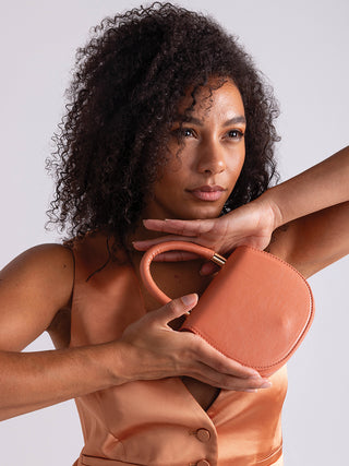 Vegan mini saddle bag in coral pink held in hands, rounded flap and top handle.